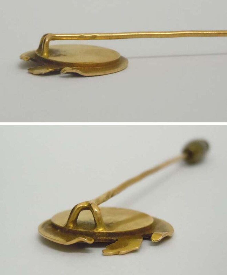 May include: A gold-toned pin with a round, slightly raised, circular design. The pin has a long, thin, straight shaft.