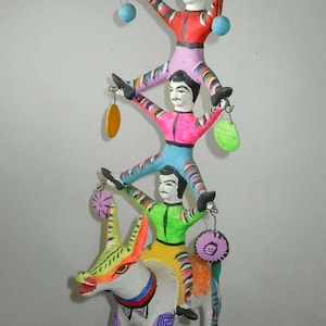 May include: A colorful folk art figurine of three figures stacked on top of each other, with the bottom figure riding a donkey. The figures are painted in bright colors, including pink, blue, green, yellow, and white. The donkey is white with green, yellow, and orange details.