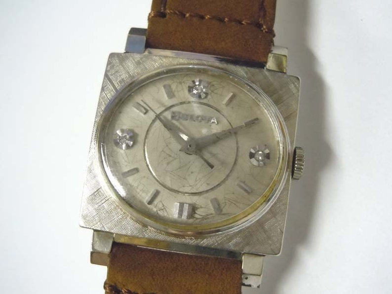 May include: A silver square-shaped wristwatch with a brown leather band. The watch face has a silver background with black Roman numeral hour markers and a silver second hand. There are three small diamonds on the watch face.