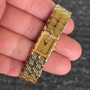 May include: A gold-toned Bulova watch with a rectangular face and a diamond accent. The watch features a gold band with a woven design. The watch face displays the Bulova logo. The watch is held in a hand.