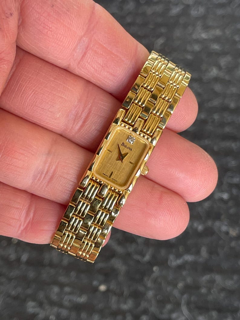 May include: A gold-toned Bulova watch with a rectangular face and a diamond accent. The watch features a gold band with a woven design. The watch face displays the Bulova logo. The watch is held in a hand.