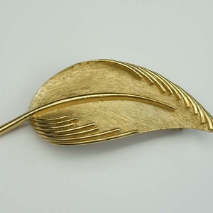 May include: A gold-toned leaf-shaped brooch with a textured surface and a raised line down the center.