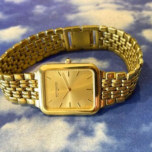 May include: A gold-toned Wittnauer watch with a rectangular face and a matching gold link band. The watch face has a gold dial with gold hands and hour markers. The watch is displayed on a blue and white patterned surface.
