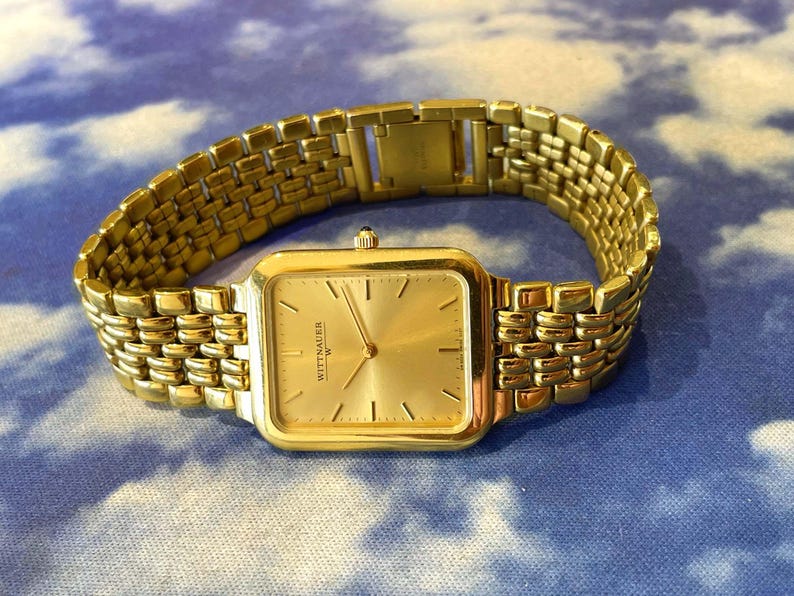 May include: A gold-toned Wittnauer watch with a rectangular face and a matching gold link band. The watch face has a gold dial with gold hands and hour markers. The watch is displayed on a blue and white patterned surface.