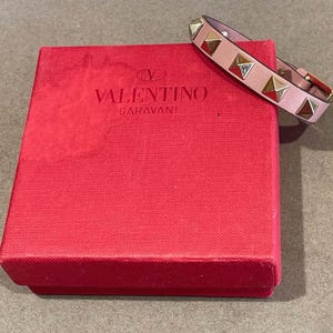 Puede incluir: Un brazalete de cuero rosa con tachuelas doradas en forma de pir&aacute;mide. El brazalete est&aacute; en una caja roja con el logotipo de Valentino Garavani.