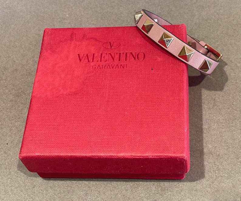 Puede incluir: Un brazalete de cuero rosa con tachuelas doradas en forma de pir&aacute;mide. El brazalete est&aacute; en una caja roja con el logotipo de Valentino Garavani.