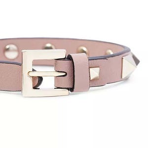 Puede incluir: Un brazalete de cuero rosa con una hebilla dorada y tachuelas piramidales plateadas.