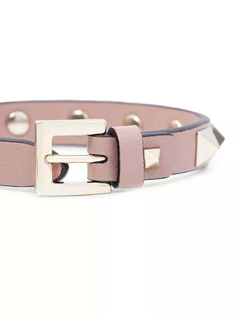 Puede incluir: Un brazalete de cuero rosa con una hebilla dorada y tachuelas piramidales plateadas.