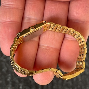 May include: A gold-toned wristwatch with a rectangular face and a matching link bracelet. The watch is held in a hand, showcasing its polished metal finish and intricate design. The watch band is approximately 6 inches in length.