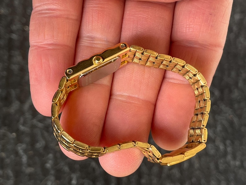 May include: A gold-toned wristwatch with a rectangular face and a matching link bracelet. The watch is held in a hand, showcasing its polished metal finish and intricate design. The watch band is approximately 6 inches in length.