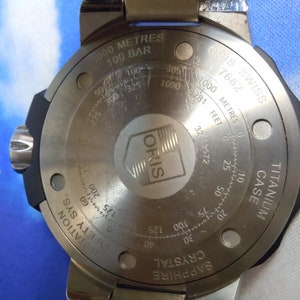 Puede incluir: La parte posterior de un reloj Oris plateado con un bisel negro. El reloj tiene un cristal de zafiro, una caja de titanio y es resistente al agua hasta 1000 metros o 100 bares. La esfera del reloj tiene el texto "ORIS SWISS 7682" y "SAPPHIRE CRYSTAL".