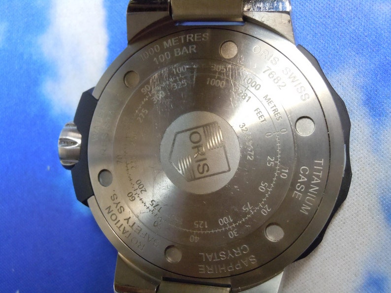 Puede incluir: La parte posterior de un reloj Oris plateado con un bisel negro. El reloj tiene un cristal de zafiro, una caja de titanio y es resistente al agua hasta 1000 metros o 100 bares. La esfera del reloj tiene el texto "ORIS SWISS 7682" y "SAPPHIRE CRYSTAL".