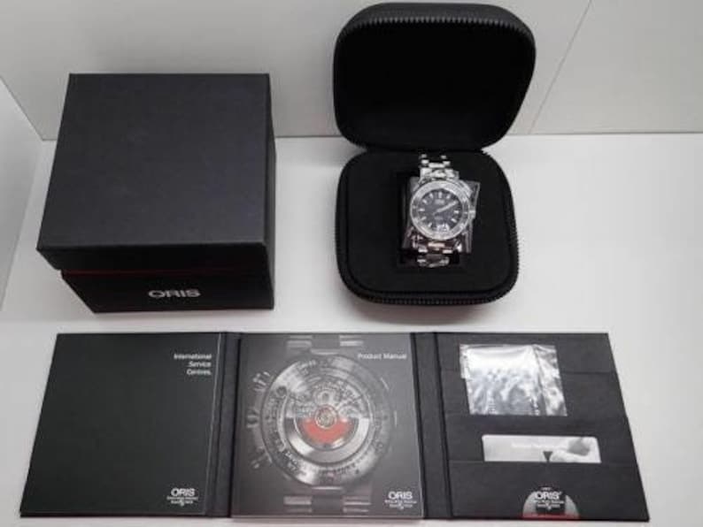 Puede incluir: Un reloj Oris plateado con una esfera negra y una ventana de fecha. El reloj est&aacute; en una caja negra con un estuche de reloj negro. El reloj viene con un manual de producto y un folleto de servicio internacional.