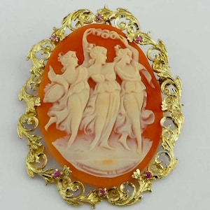 May include: A gold cameo brooch featuring a carved shell cameo of three women in a circle. The cameo is set within a gold filigree frame with pink gemstones.