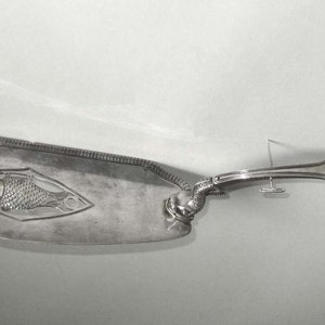 May include: A silver serving utensil with a fish design on the handle. The utensil is shaped like a long, thin spatula with a rounded end.