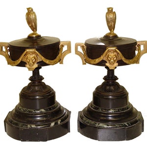 Könnte beinhalten: Ein Paar verzierte Urnen aus Bronze und Marmor mit Löwenkopf-Akzenten und Eulen-Fialen. Die Urnen sind mit goldenen Akzenten verziert und haben einen schwarzen Marmorsockel.