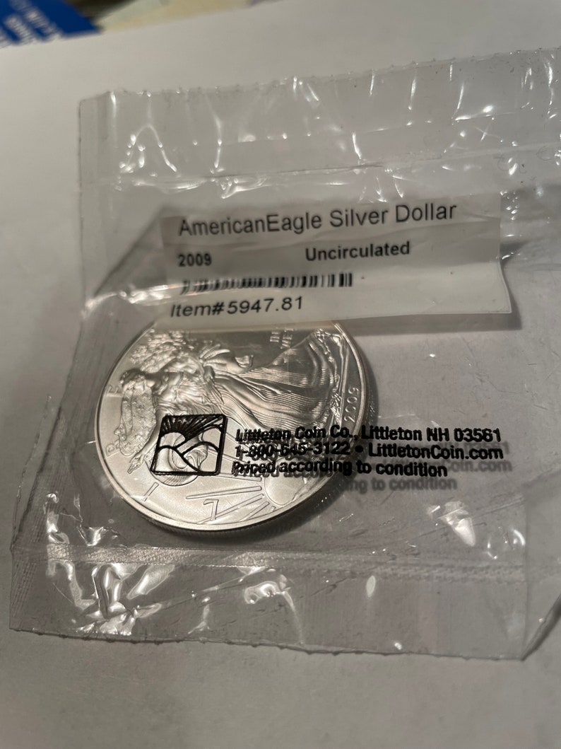 K&ouml;nnte beinhalten: Eine silberne American Eagle Dollar-M&uuml;nze in einem Plastikbeutel. Die M&uuml;nze ist mit 2009 datiert und ungezirkelt. Der Beutel tr&auml;gt ein Etikett mit der Aufschrift "Littleton Coin Co., Littleton NH 03561, 1-800-645-3122, LittletonCoin.com, Priced according to condition".