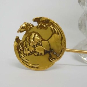 May include: A gold-toned brooch featuring a detailed eagle design with a raised profile. The eagle is depicted with its wings spread and head turned to the side. The brooch has a pin on the back for attaching to clothing.