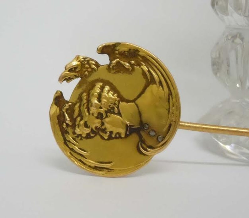 May include: A gold-toned brooch featuring a detailed eagle design with a raised profile. The eagle is depicted with its wings spread and head turned to the side. The brooch has a pin on the back for attaching to clothing.