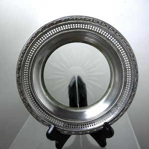 May include: A silver-colored, round decorative tray with an etched glass center featuring a sunburst design. The tray has a detailed, patterned border and is supported by a black stand. The tray is likely vintage or antique.