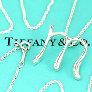 May include: A silver necklace with a stylized letter "M" pendant. The pendant has a smooth, polished finish and hangs from a delicate chain. The background is a light blue color, and the text "TIFFANY & Co." is visible.