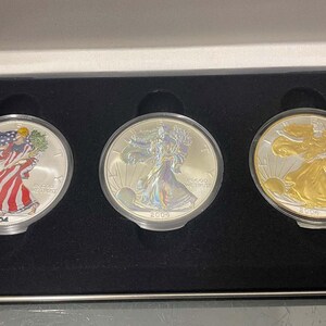 Puede incluir: Tres monedas de plata American Eagle en una vitrina. Las monedas son de los años 2004, 2005 y 2006. Las monedas presentan un diseño de la Dama de la Libertad caminando con una antorcha y una rama de olivo.