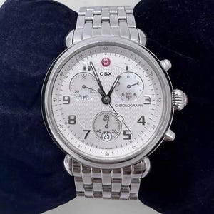 May include: A silver chronograph wristwatch with a white face and black markings. The watch has three subdials, one for seconds, one for minutes, and one for 1/10 of a second. The watch face also has the words "CSX" and "CHRONOGRAPH" printed on it.