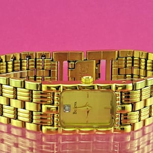May include: A gold-toned Bulova watch with a rectangular face and a matching gold bracelet. The watch face has gold hands and a small diamond accent. The bracelet is made of interlocking rectangular links.