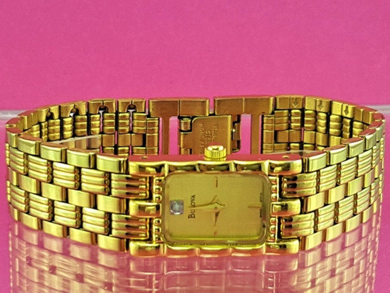 May include: A gold-toned Bulova watch with a rectangular face and a matching gold bracelet. The watch face has gold hands and a small diamond accent. The bracelet is made of interlocking rectangular links.