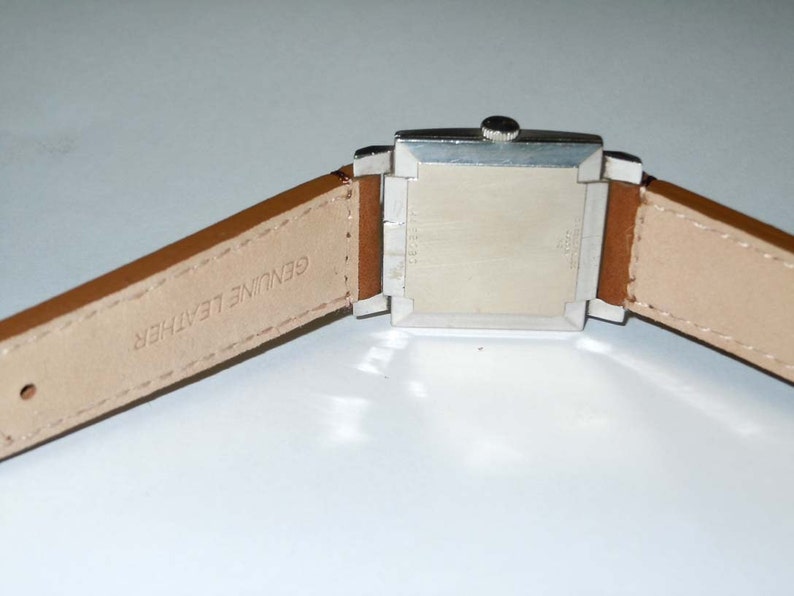 May include: A silver square watch with a brown leather band. The band is labeled "Genuine Leather".