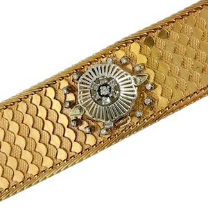 May include: A gold bracelet with a decorative center piece featuring a cluster of diamonds. The bracelet is made of a woven, fish scale pattern.