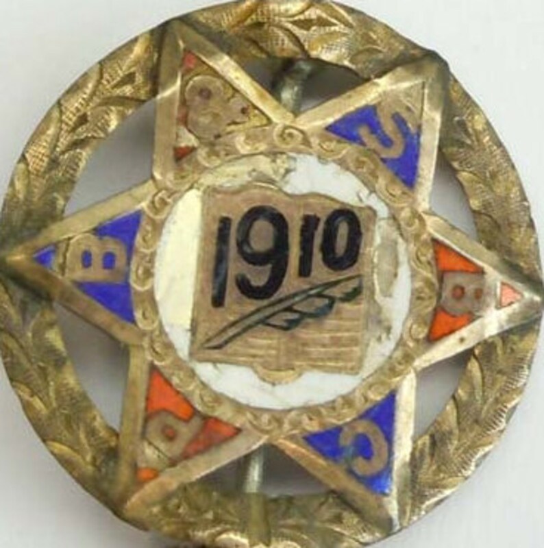 K&ouml;nnte beinhalten: Eine sternf&ouml;rmige Anstecknadel in Goldton mit blauem, rotem und wei&szlig;em Email. Die Mitte des Sterns zeigt ein Buch mit dem Jahr "1910" und einer Feder.