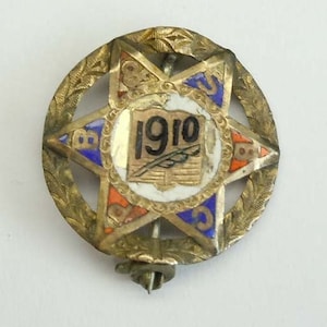 K&ouml;nnte beinhalten: Eine goldfarbene sternf&ouml;rmige Anstecknadel mit blauem, rotem und wei&szlig;em Email. In der Mitte des Sterns befindet sich ein Buch mit einer Feder und dem Jahr "1910".