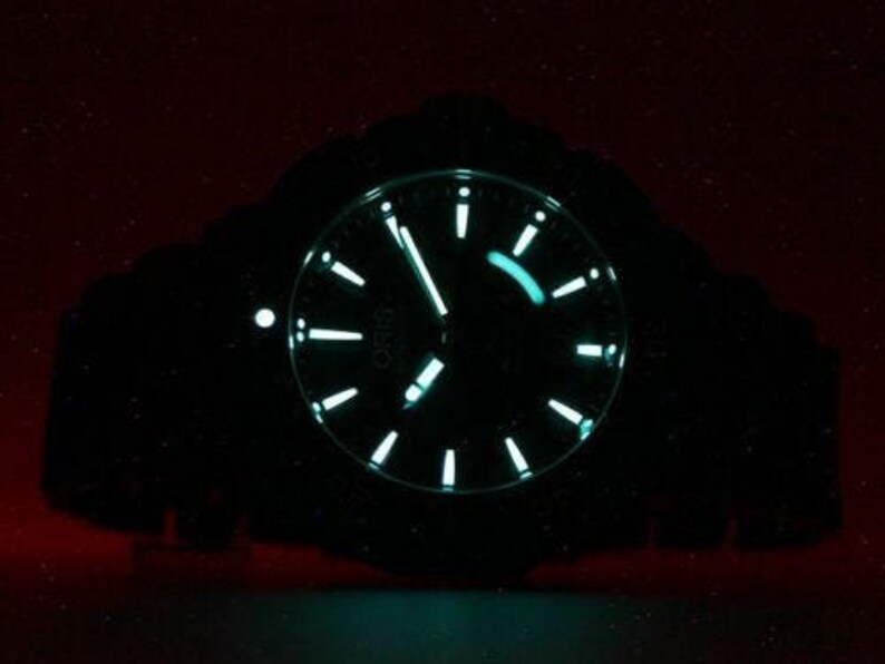 Puede incluir: Un reloj de pulsera negro con una esfera verde luminosa y agujas plateadas. La esfera del reloj tiene una ventana de fecha y un segundero. El reloj est&aacute; iluminado en la oscuridad.
