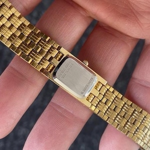 May include: A gold-toned watch with a rectangular face and a woven band. The watch band is made of small, rectangular links. The watch face is silver-colored. The watch is held in a hand.