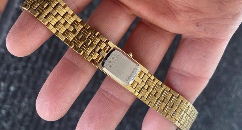 May include: A gold-toned watch with a rectangular face and a woven band. The watch band is made of small, rectangular links. The watch face is silver-colored. The watch is held in a hand.