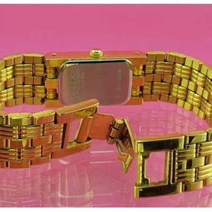 May include: A gold-toned wristwatch with a rectangular face and a woven-style band. The watch is shown from the back, with the clasp open. The watch band appears to be approximately 0.75 inches wide.