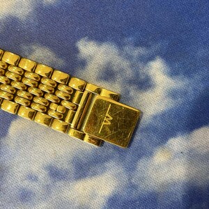 May include: Close-up of a gold-toned watch band with a rectangular clasp. The band features a series of small, rounded rectangular links. The clasp is engraved with the letters "TW". The background is a blue and white cloudy pattern.
