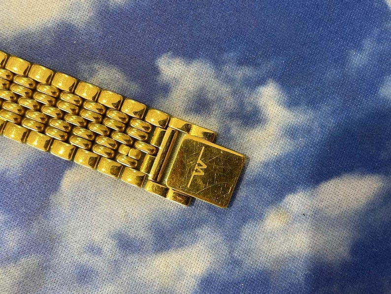 May include: Close-up of a gold-toned watch band with a rectangular clasp. The band features a series of small, rounded rectangular links. The clasp is engraved with the letters "TW". The background is a blue and white cloudy pattern.