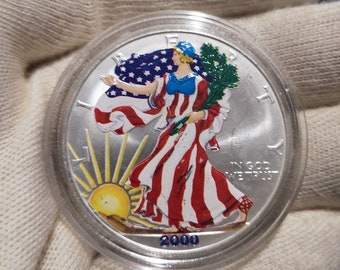 May include: A silver American Eagle coin with a colorized design of Lady Liberty holding a branch of olive leaves and a flag. The coin is dated 2000 and has the inscription "In God We Trust".