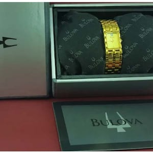 May include: A gold-toned Bulova watch with a rectangular face and a woven band, presented in a silver box. The watch is displayed on a black cushion inside the box, with the Bulova logo visible. The watch is a classic timepiece.