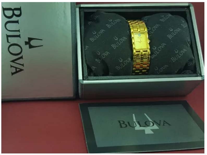 May include: A gold-toned Bulova watch with a rectangular face and a woven band, presented in a silver box. The watch is displayed on a black cushion inside the box, with the Bulova logo visible. The watch is a classic timepiece.