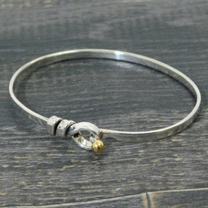 May include: A silver bangle bracelet with a gold accent. The bracelet has a unique clasp that is shaped like a knot.