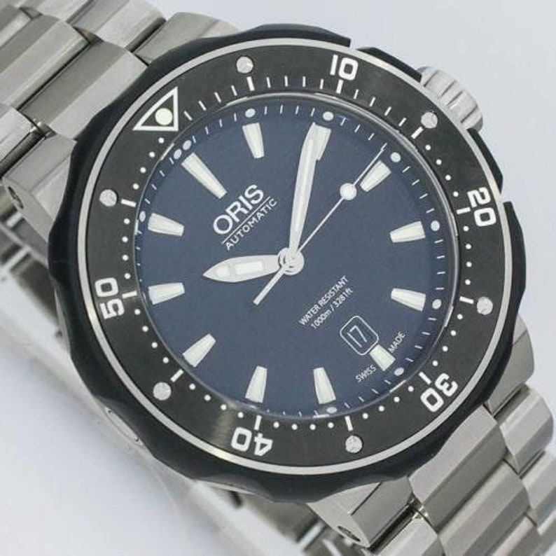 Puede incluir: Un reloj de pulsera Oris para hombre de color negro y plateado con una esfera azul y agujas plateadas. El reloj tiene un bisel negro con marcas blancas y el texto "WATER RESISTANT 1000m / 3281ft" y "SWISS MADE".