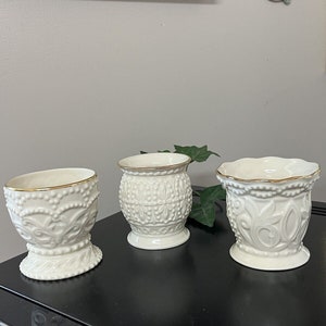 May include: Three off-white ceramic vases with gold trim. The vases have ornate embossed designs, including floral and beaded patterns. They are displayed on a black surface, with a hint of greenery in the background. The vases vary slightly in shape and size.