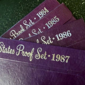 May include: A set of five purple envelopes with silver lettering. The envelopes are labeled "Proof Set" and the years 1984, 1985, 1986, and 1987.