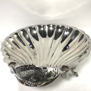 May include: A silver-colored, shell-shaped dish with a detailed fish sculpture at the base. The dish has a scalloped edge and two small, cylindrical holders. The fish is intricately designed, adding a decorative element to the piece.