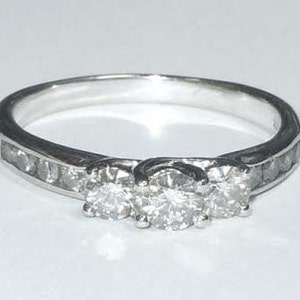 May include: A silver ring with three round diamonds set in a band with smaller diamonds. The ring is on a white background.
