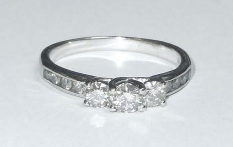 May include: A silver ring with three round diamonds set in a band with smaller diamonds. The ring is on a white background.