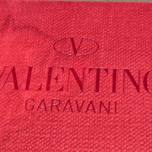 Puede incluir: Etiqueta de cuero roja con el texto "VALENTINO GARAVANI" en letras blancas y un logotipo "V" blanco en forma ovalada.